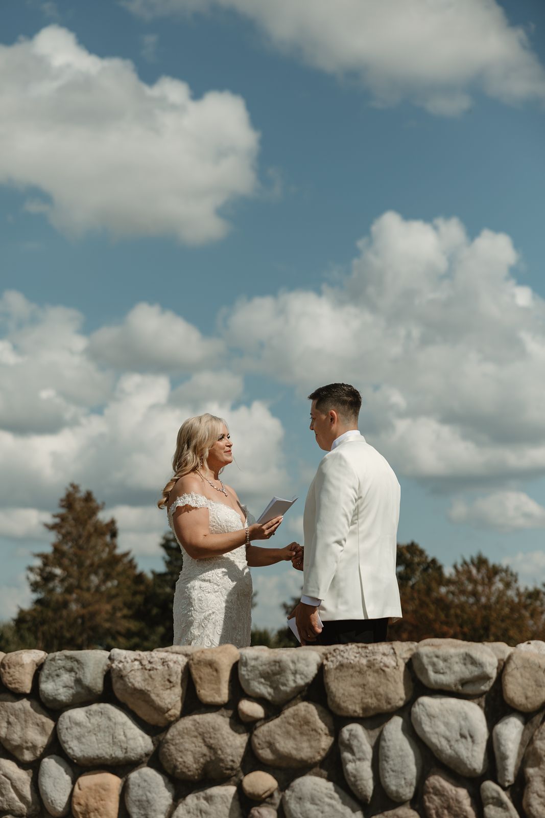 A couple exchanges vows while standing on a rustic stone wall under blue skies.