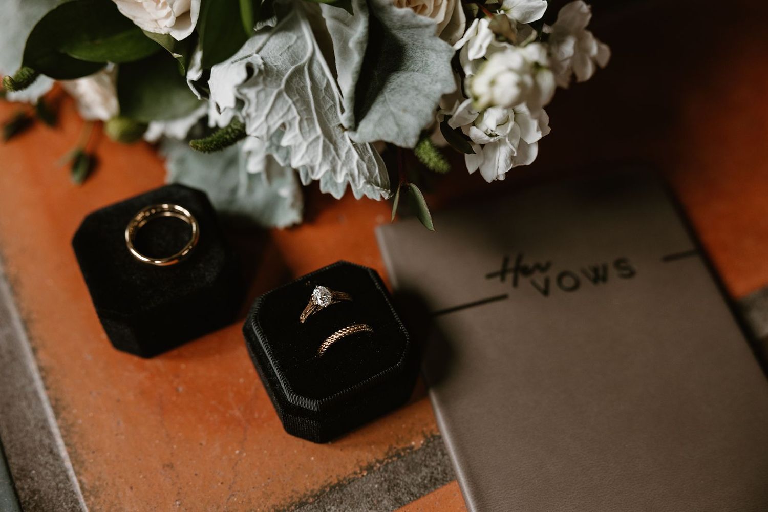 Wedding rings displayed in black boxes next to white flowers on rustic surface.