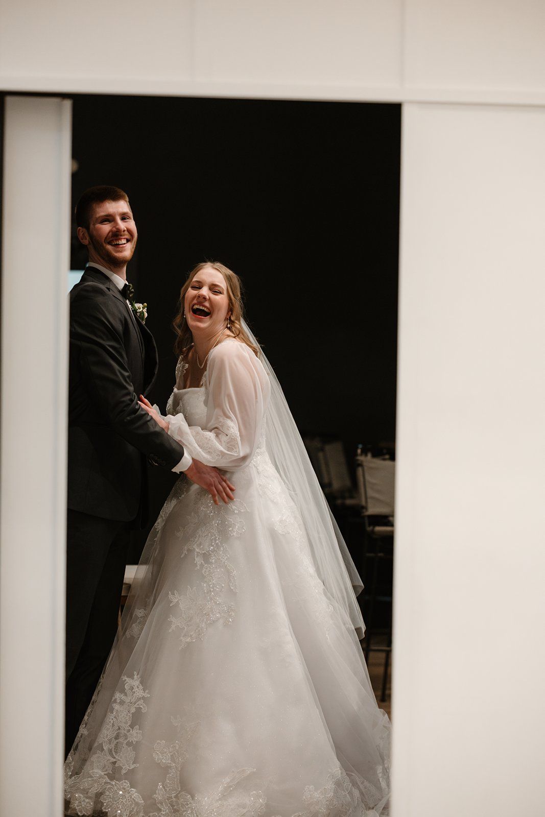 A bride and groom share a joyful candid moment in their wedding attire against a dark background.