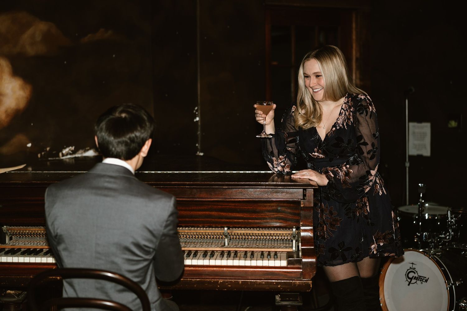Jazz musicians perform in a dimly lit lounge with a piano and drum set while a vocalist holds a drink.