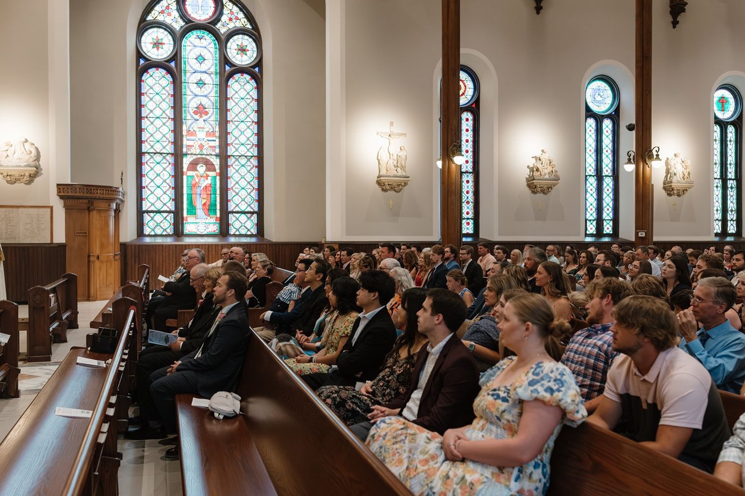 Wedding guests seated in elegant church pews with stained glass windows and ornate wall sconces.