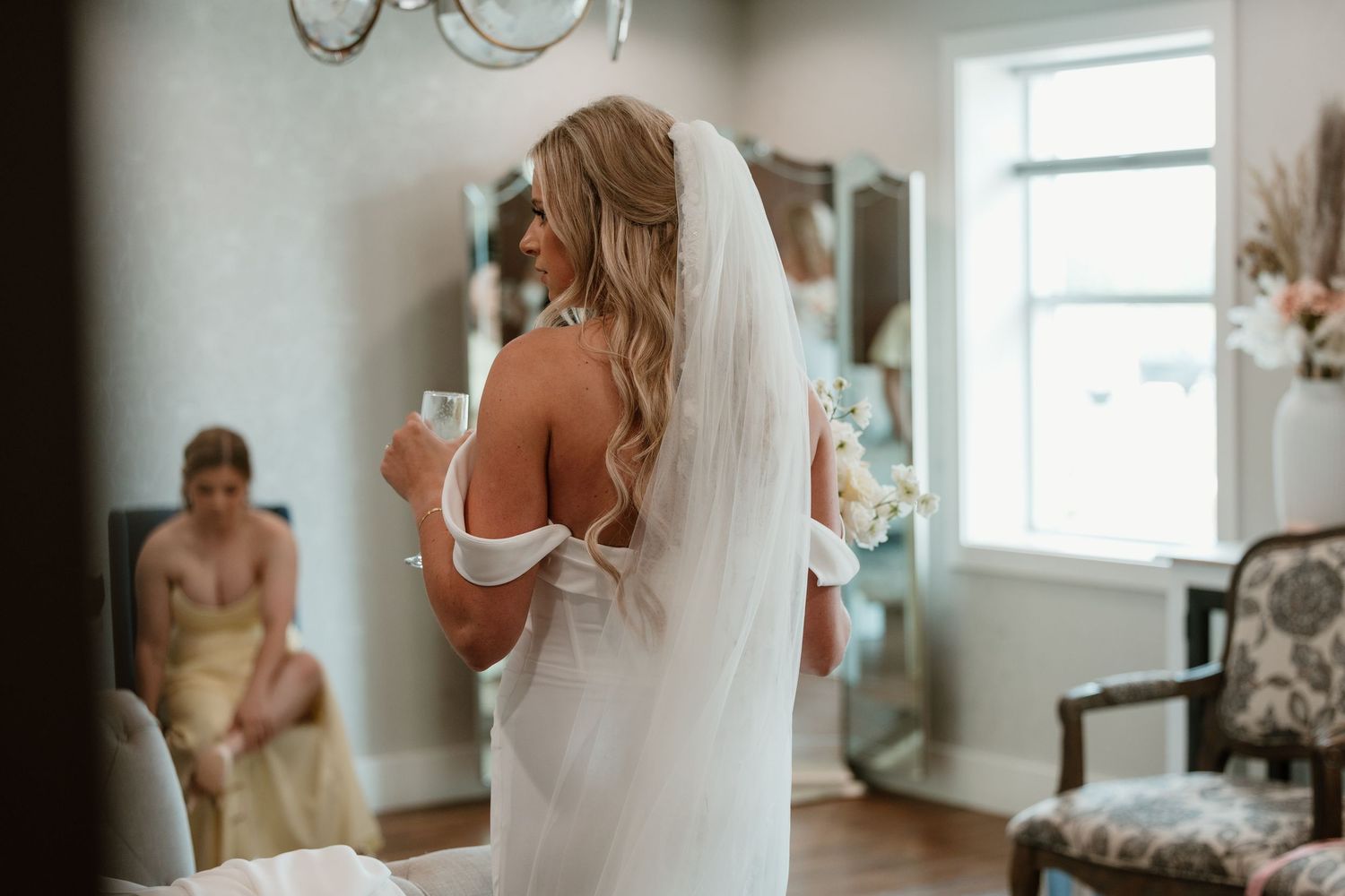 A bride in a white wedding dress gets ready in a sunlit room while looking in a mirror.