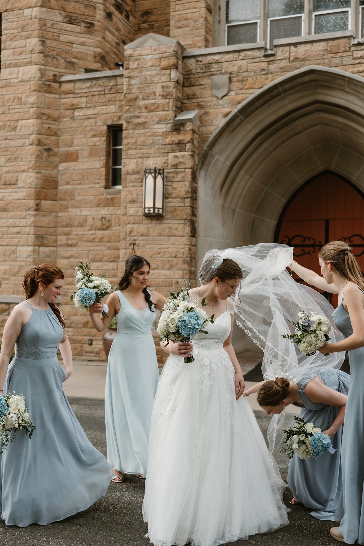 A joyful bridal party in light blue bridesmaid dresses helps adjust the bride's flowing veil outside a stone church.