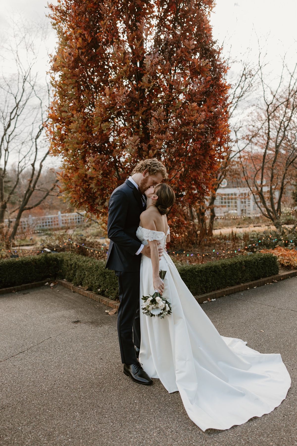 Bride and groom share a romantic kiss outdoors in autumn, with vibrant red foliage and a flowing white wedding gown.