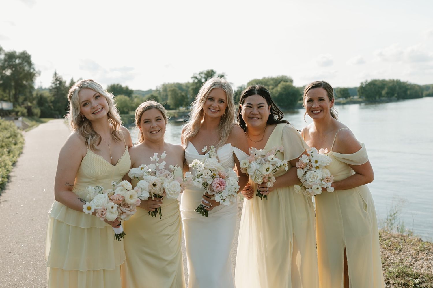Bride and four bridesmaids in yellow dresses holding white bouquets pose by a lakeside path.