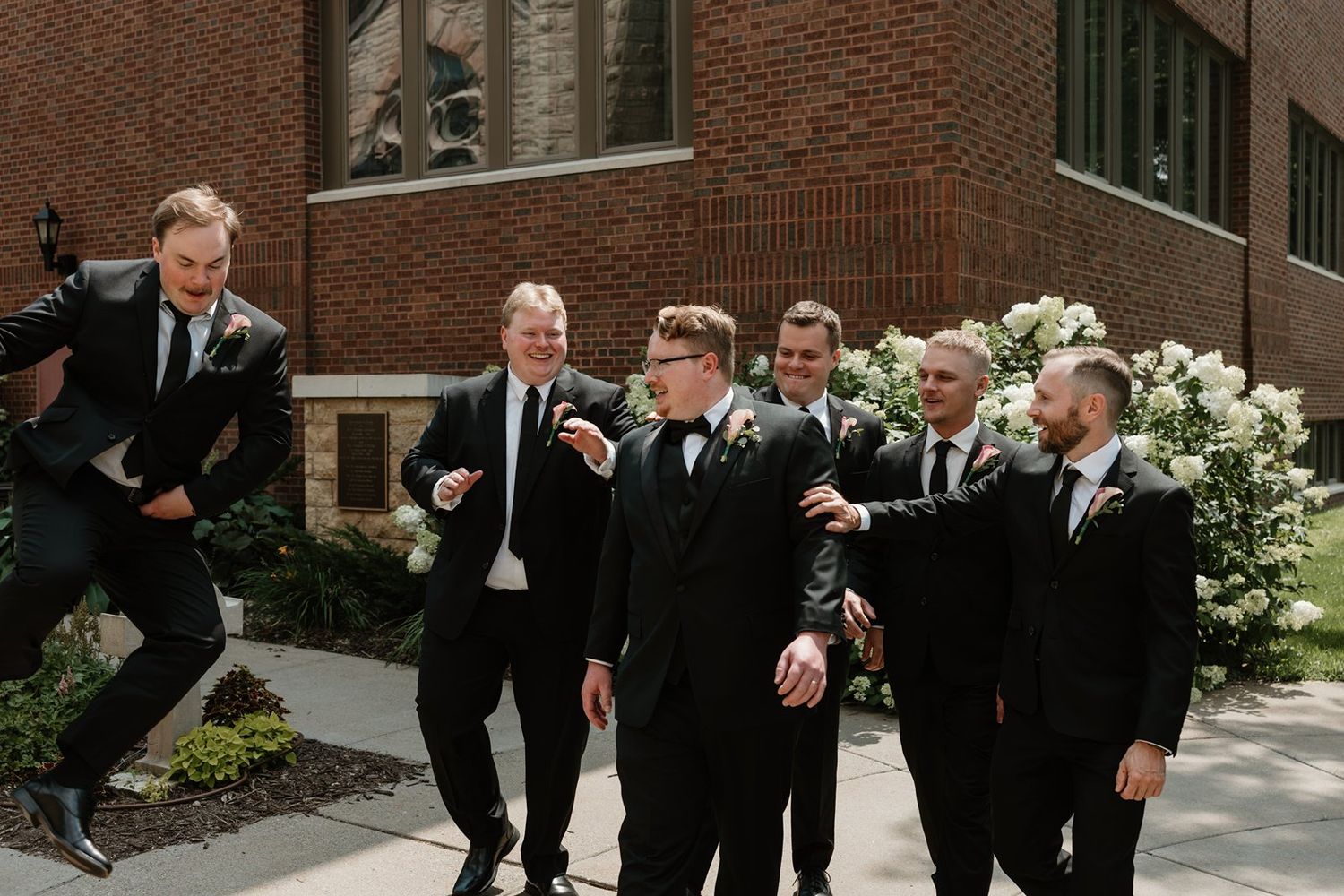 Wedding party members in dark suits pose playfully in a sequence of candid shots against a brick building.