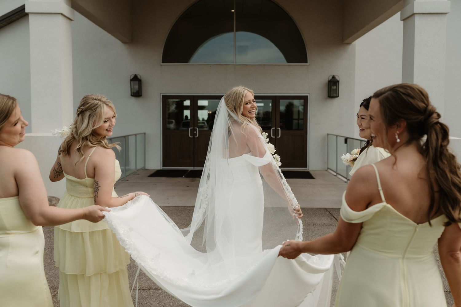 Bridesmaids in pale yellow dresses help arrange a flowing white wedding veil outside a church entrance.