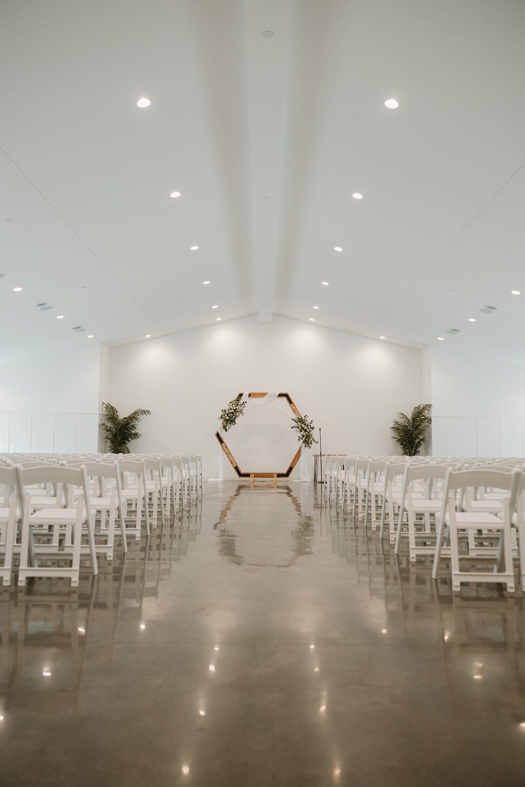 Modern minimalist wedding ceremony setup with hexagonal arch and white chairs in a sleek indoor venue with polished concrete floors.