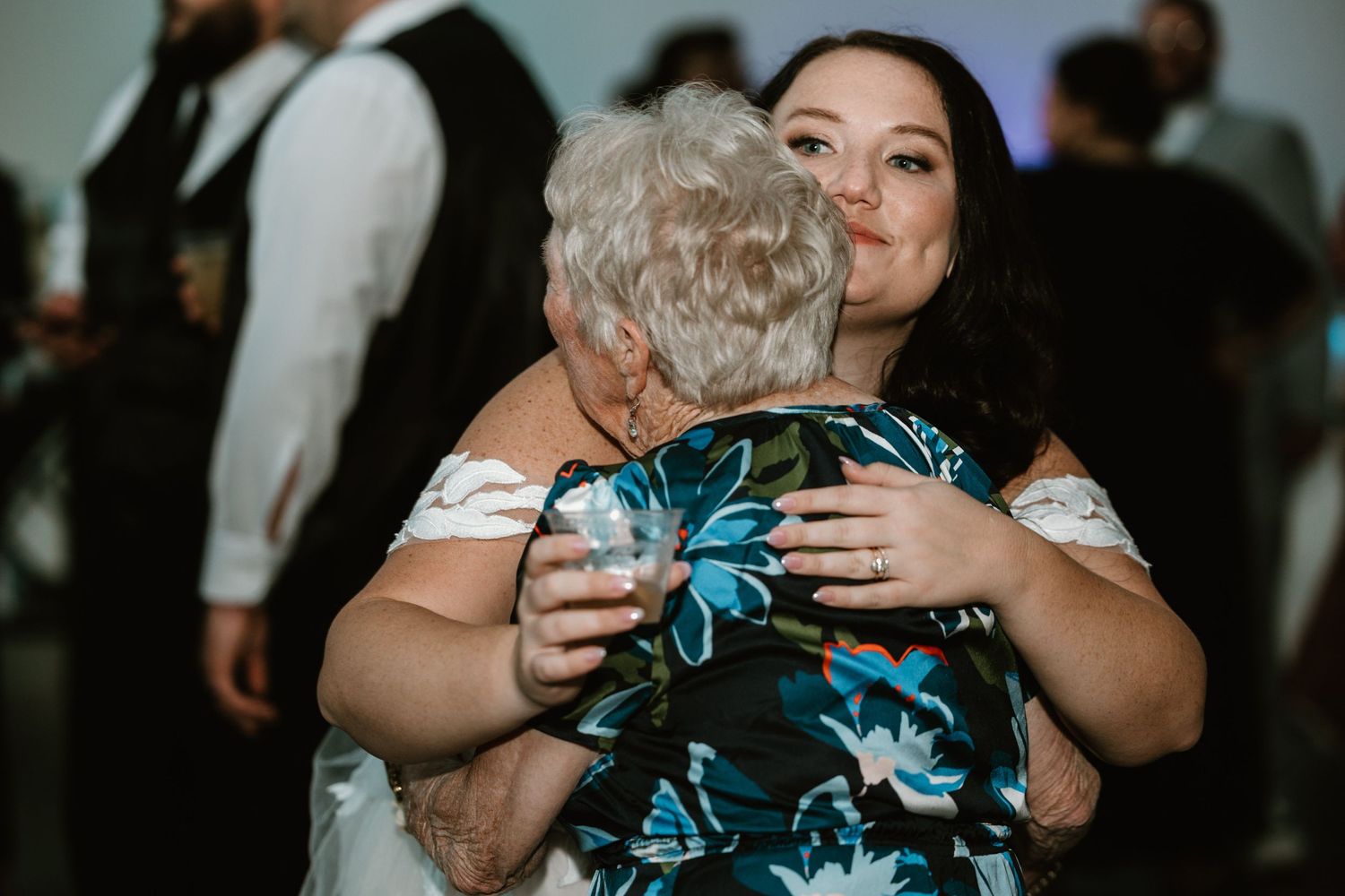 Two people share a warm embrace on the dance floor, with one wearing a floral print dress at what appears to be a celebration.