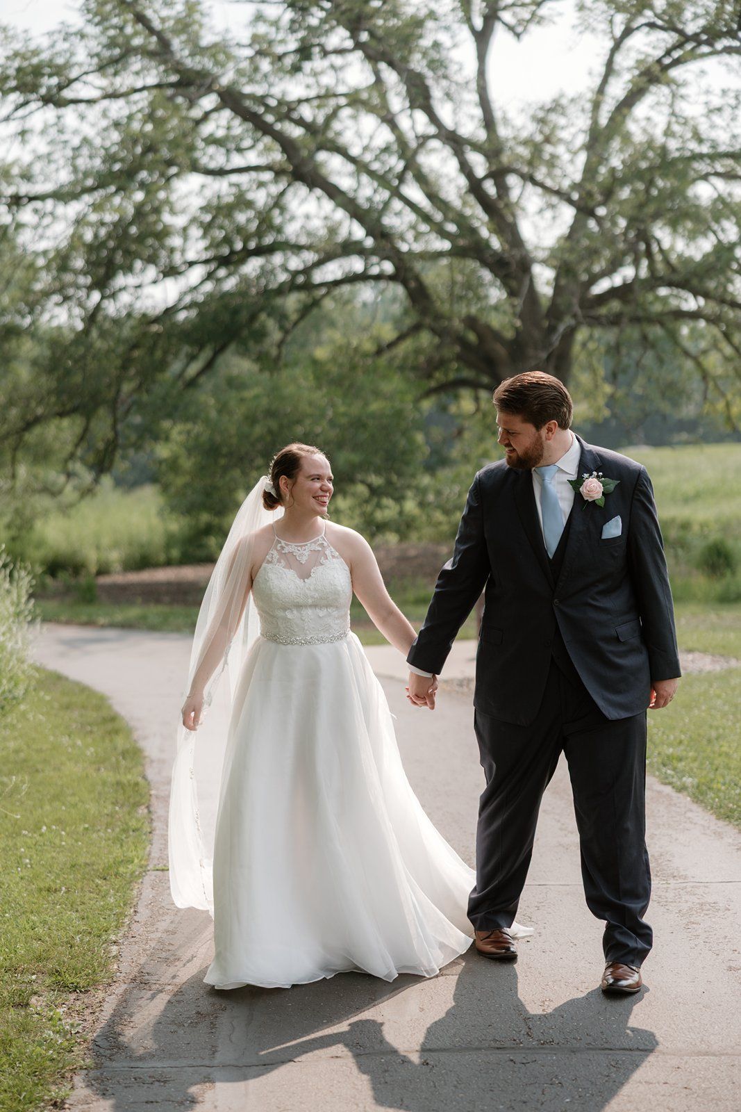 A couple in wedding attire walks together on a country path surrounded by green trees and grass.