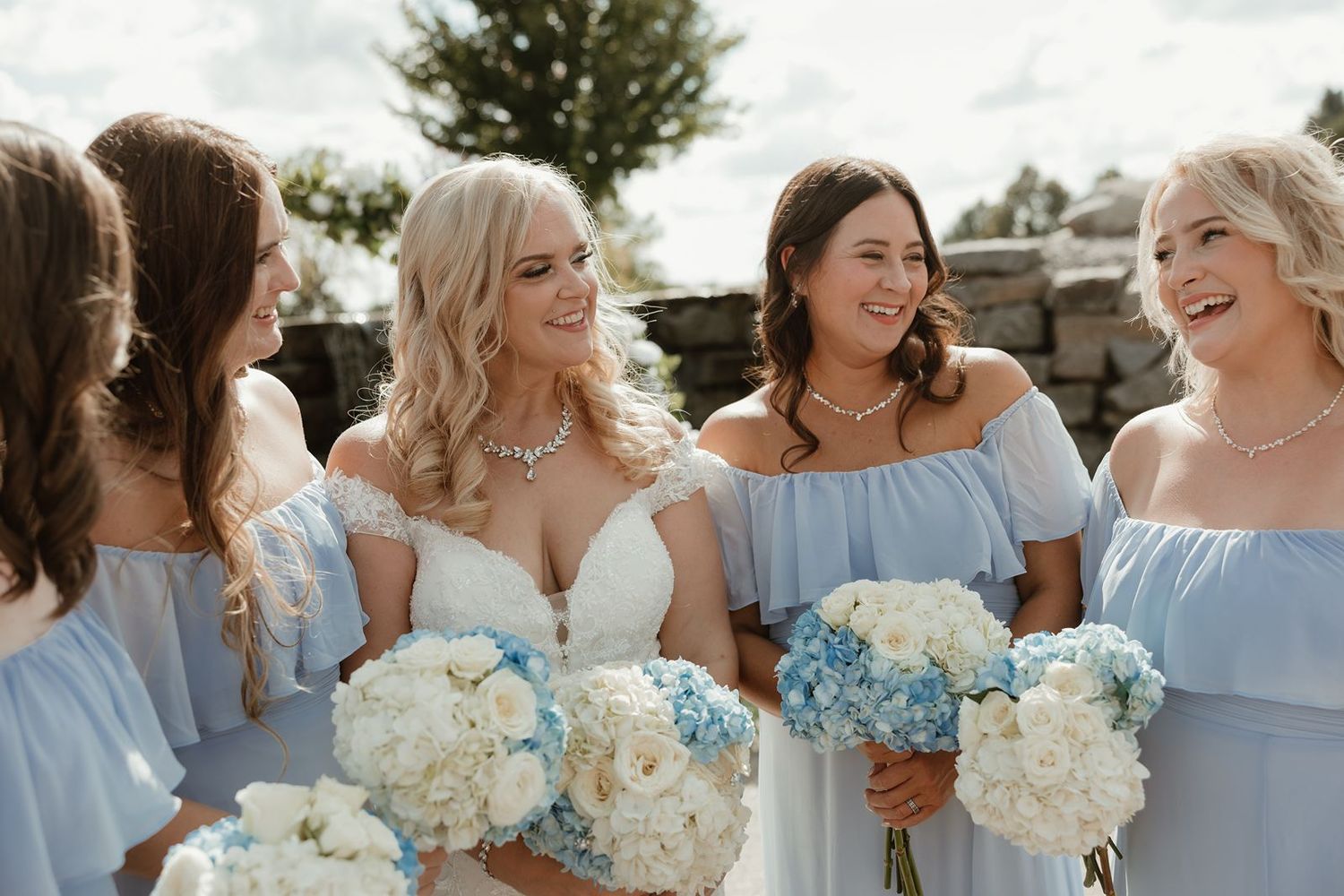 A bridal party in light blue dresses holds white and blue hydrangea bouquets while sharing a candid moment outdoors.