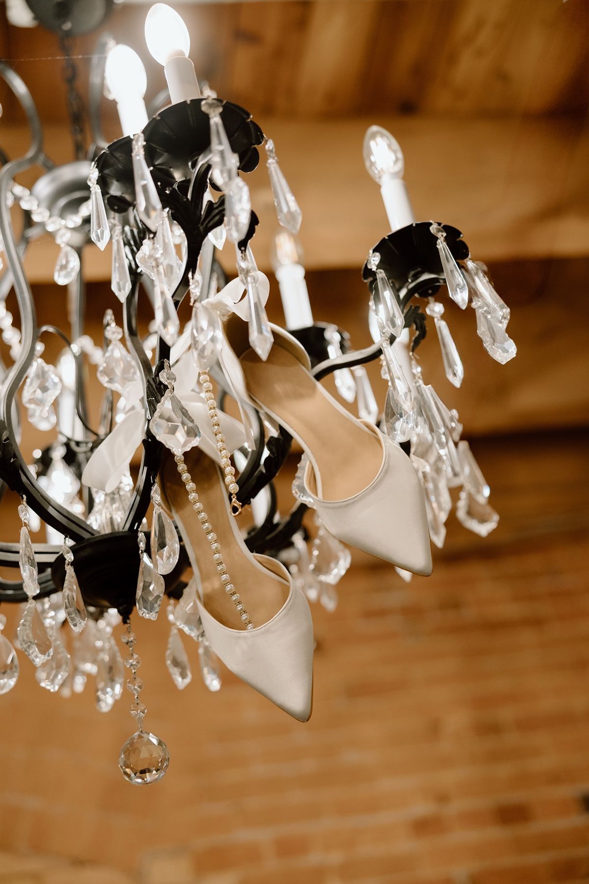 Elegant white bridal heels hanging on a black crystal chandelier in a rustic wooden venue setting.