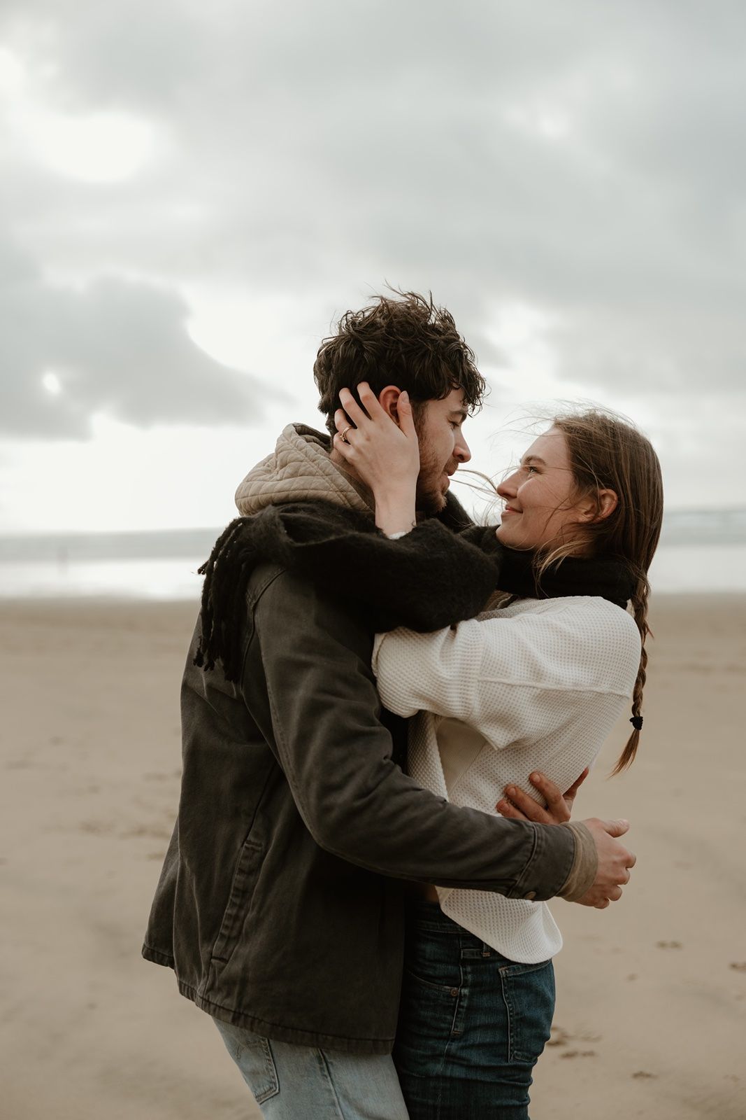 Couple embracing and smiling on a cloudy beach, wearing casual jackets, sharing a romantic moment by the ocean.