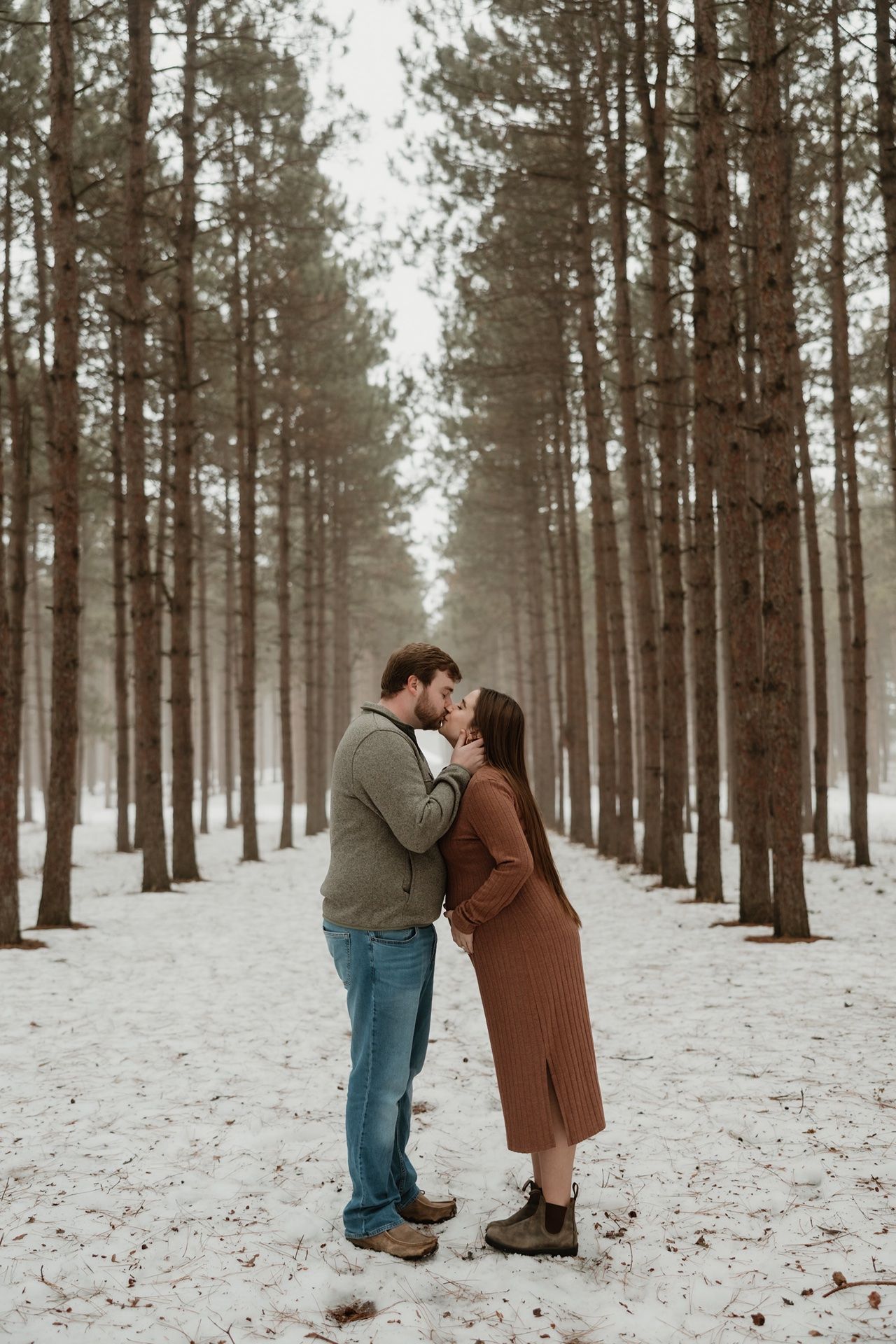 Couple sharing a romantic kiss in a snowy pine forest during winter, dressed in warm coats and casual attire.