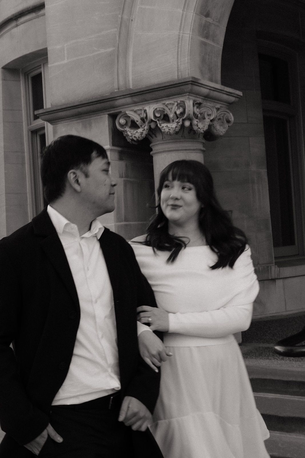 Black and white photo of a couple in formal attire standing before a classical stone building with ornate columns.