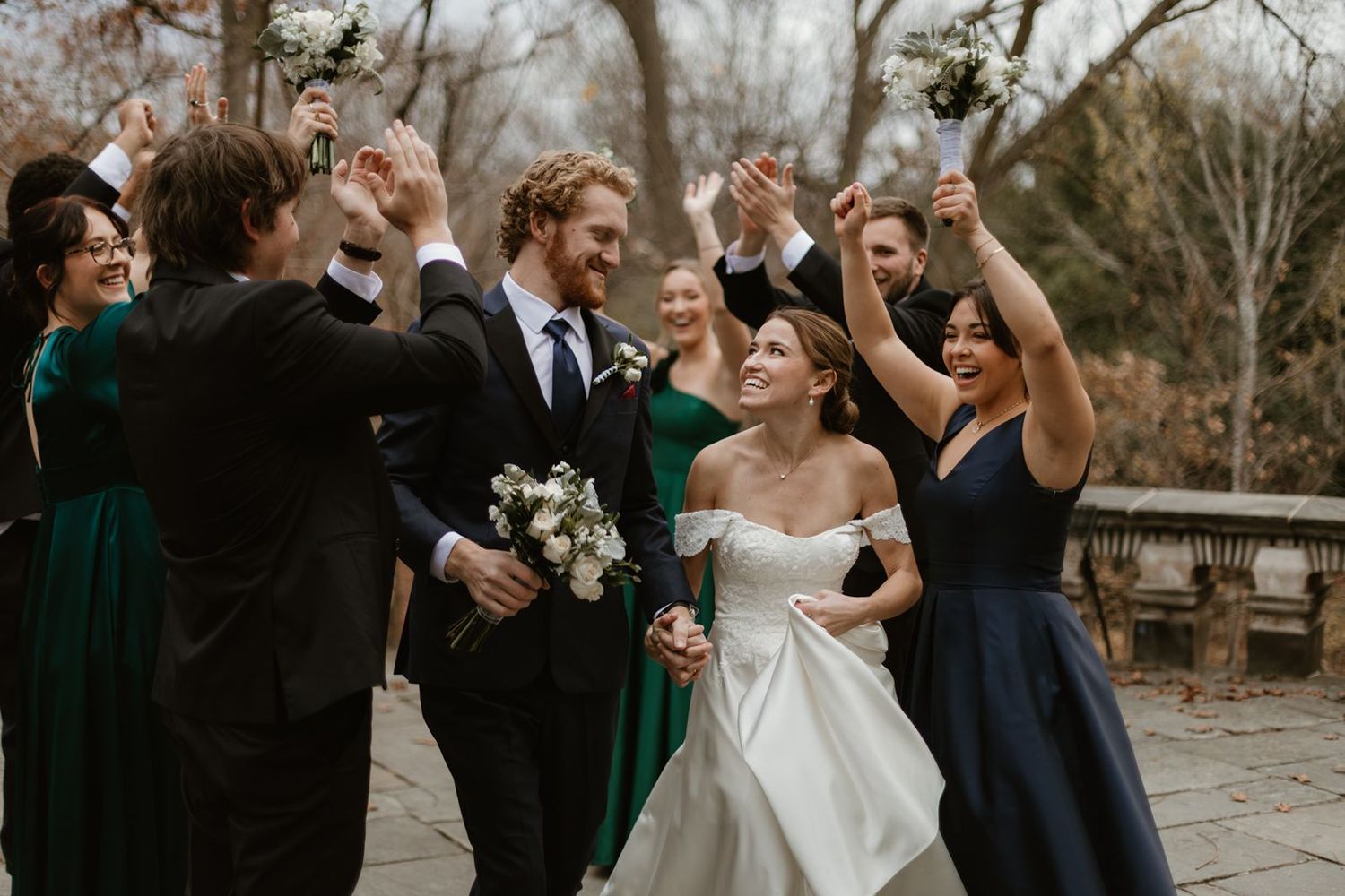 Wedding party celebrates joyfully outdoors with raised arms in navy and emerald dresses.