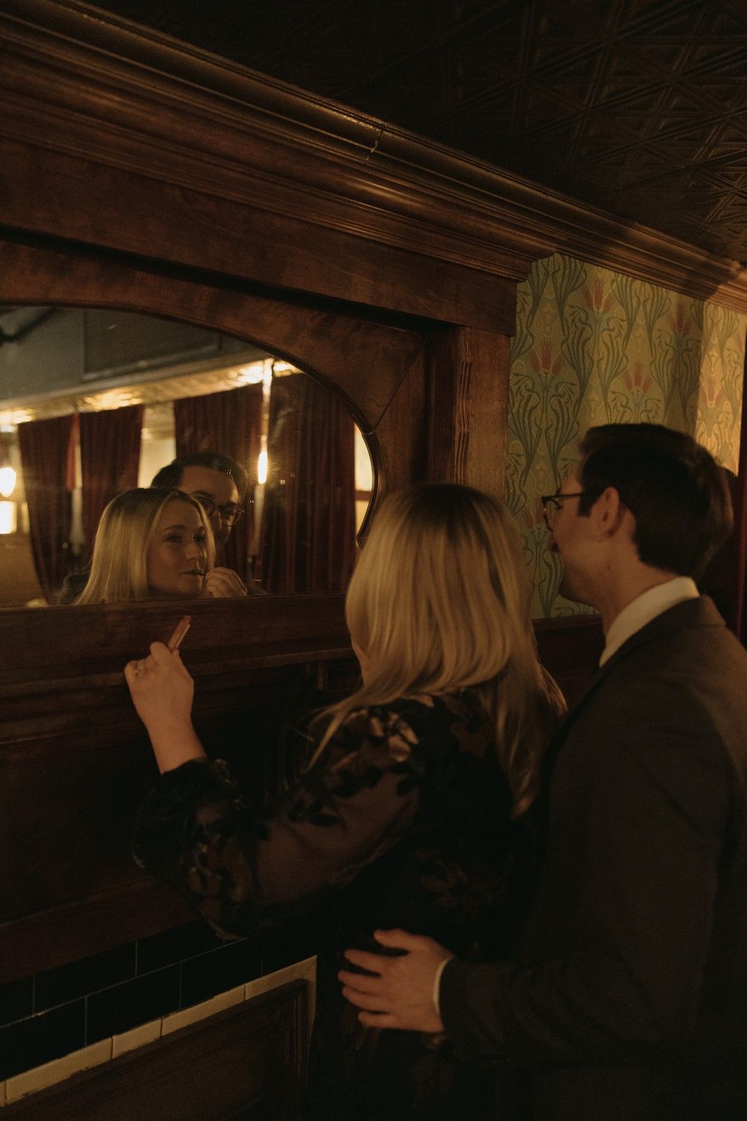 People gather at a dimly lit bar with vintage wood paneling and decorative wallpaper in a moody atmosphere.
