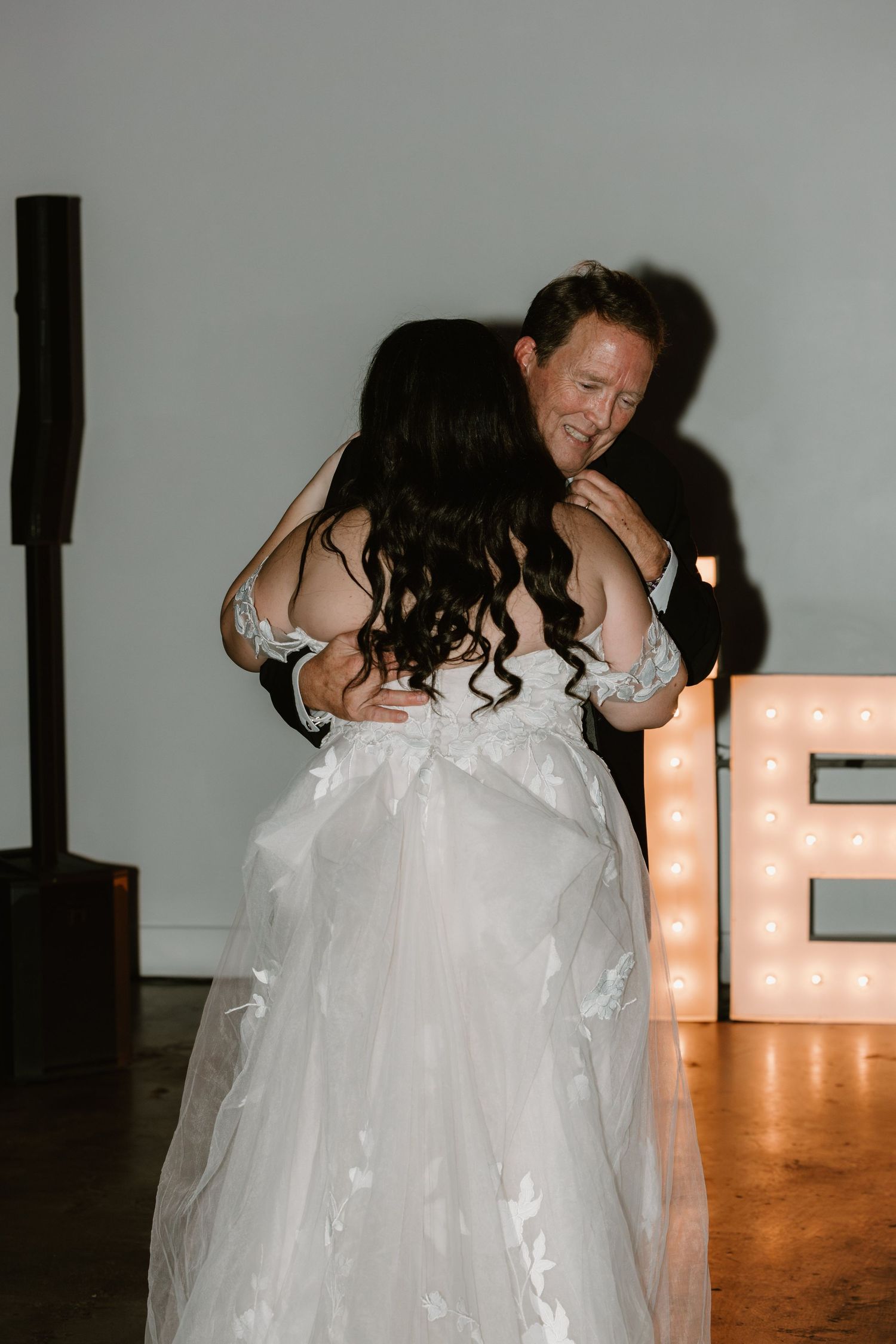 A couple embraces on the dance floor at their wedding reception, with illuminated marquee letters visible in the background.