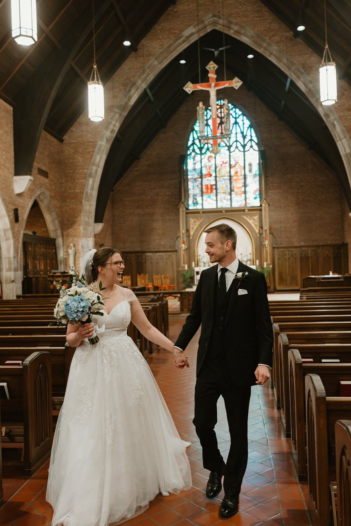 A couple walks down the aisle of a stunning historic church with stained glass windows and gothic arches.