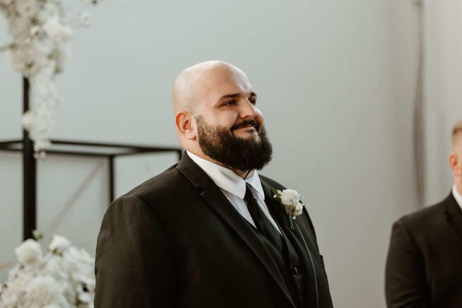 A groom in a black suit with a white boutonniere smiles joyfully during a wedding ceremony.