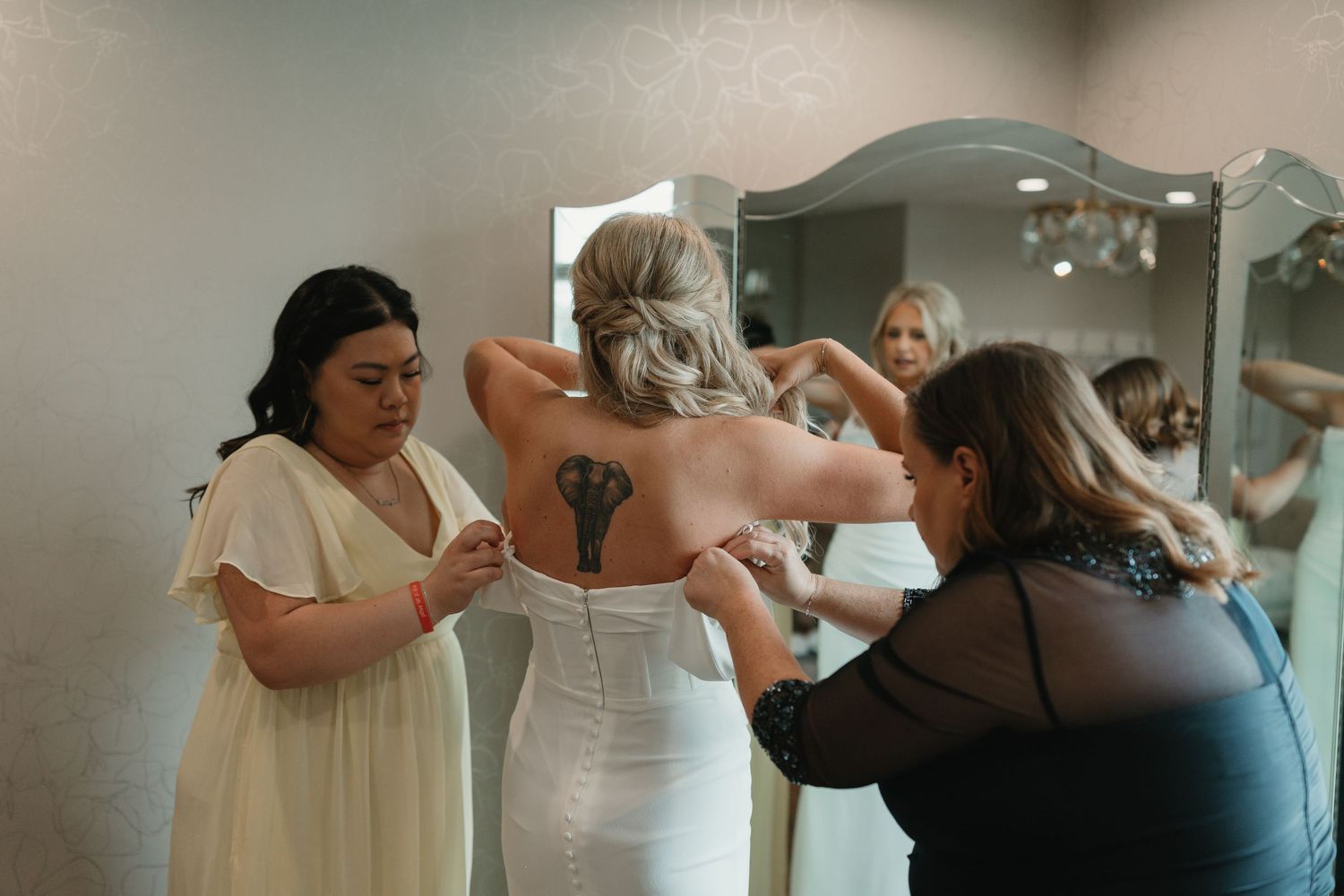Wedding party members help button up a white wedding dress in a sunlit room during preparation.