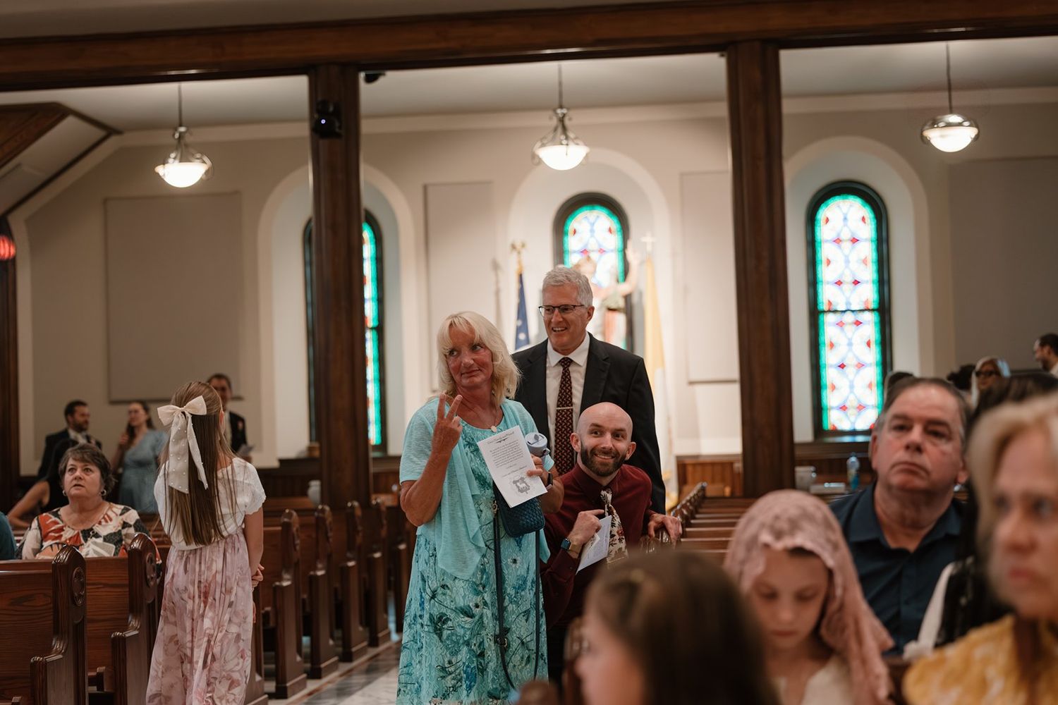Wedding guests stand and participate in a ceremony inside a church with stained glass windows and wooden pews.