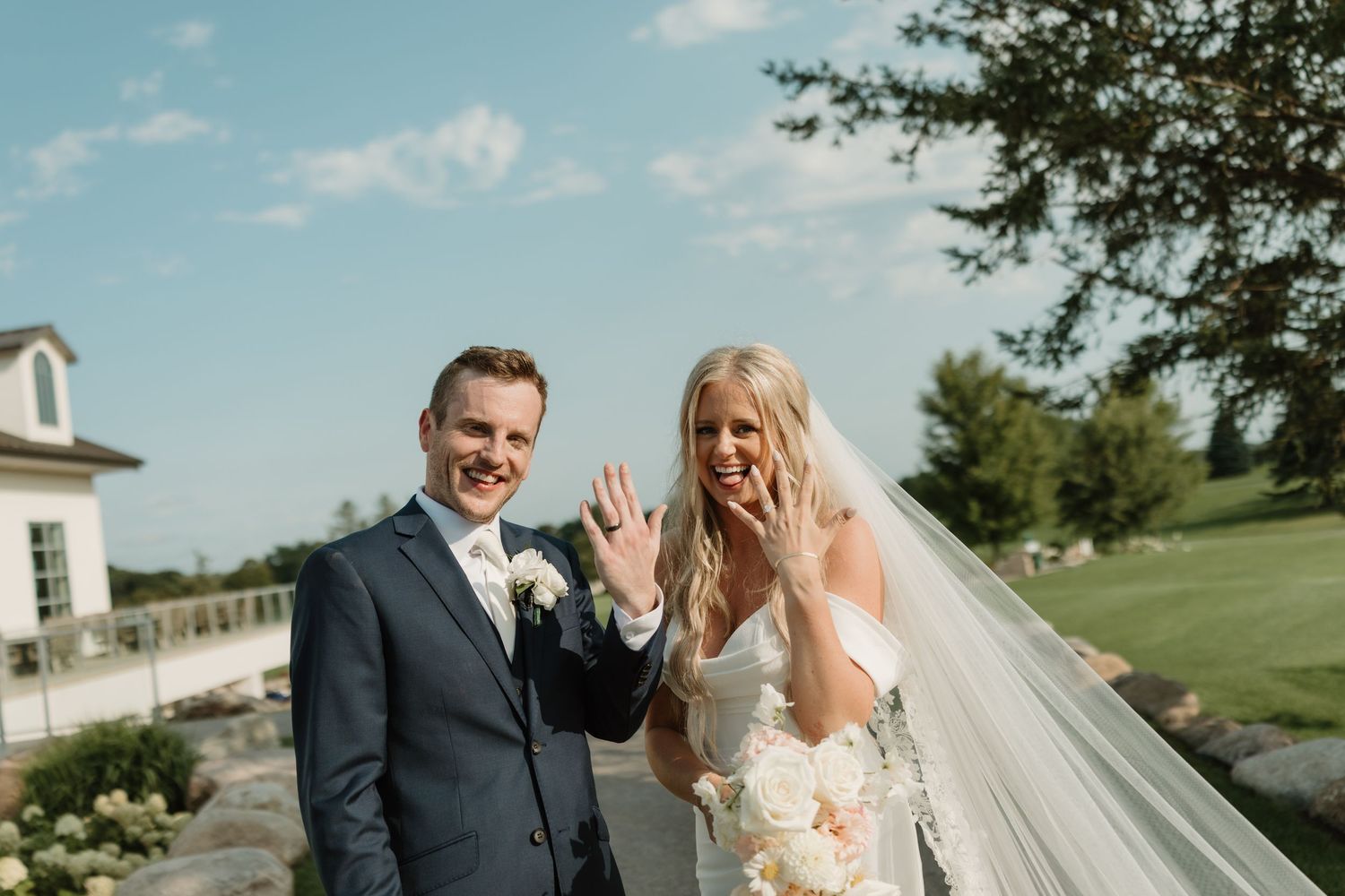A joyful wedding couple shares a candid moment outdoors on their special day, with a white building in the background.