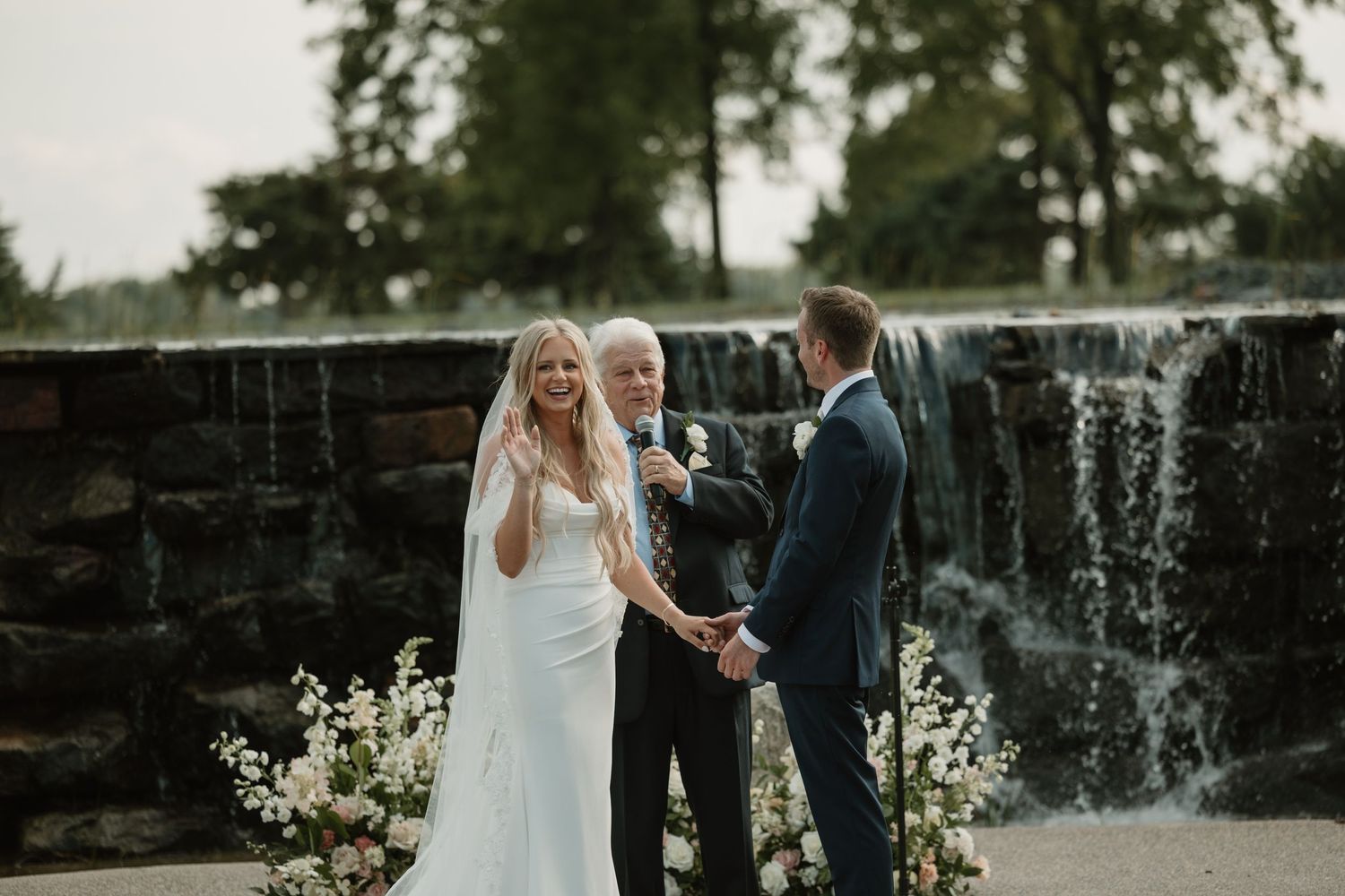 A couple in wedding attire stands near a cascading waterfall during their outdoor ceremony.