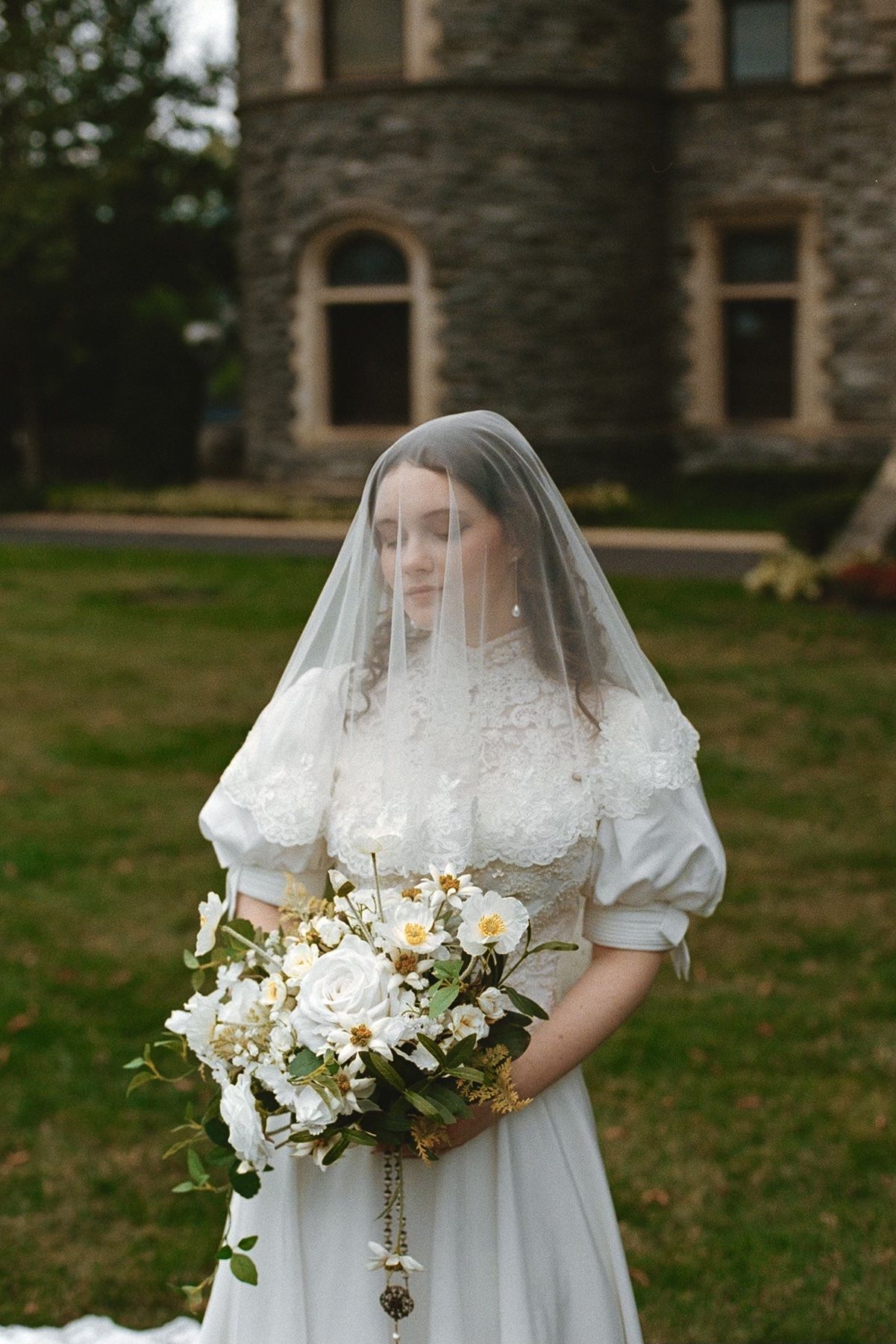 Bride in white lace dress and veil holding bouquet of white flowers outside stone building.