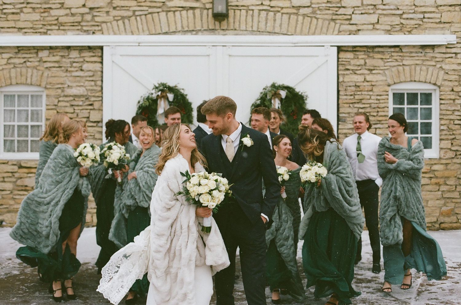 Bride and groom share a kiss surrounded by wedding party in elegant attire outside a stone building.