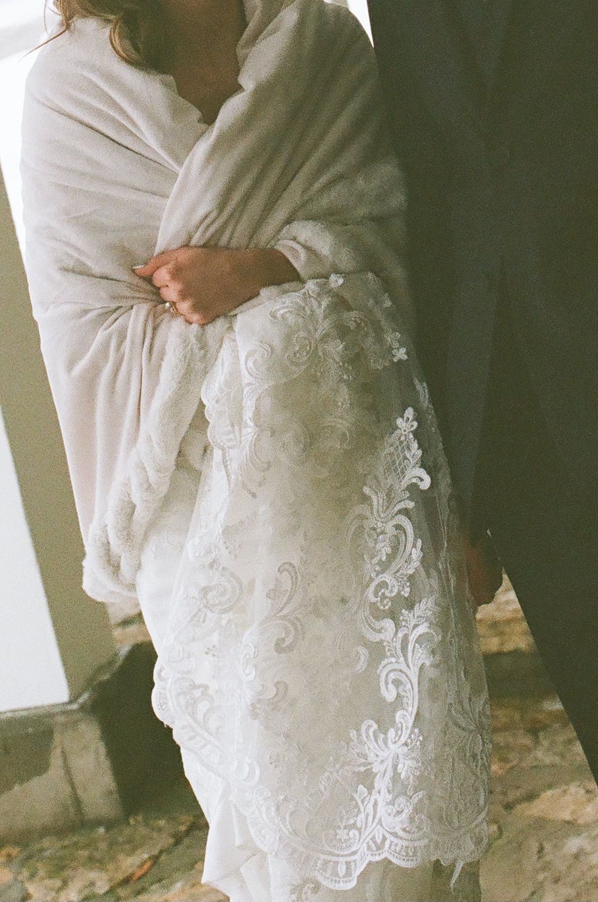 Bride in white lace wedding dress and ivory shawl embracing groom in elegant wedding ceremony photo.