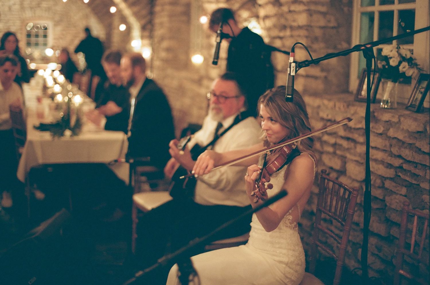Bride playing violin with guitarist at elegant rustic wedding reception with warm string lights and stone walls.