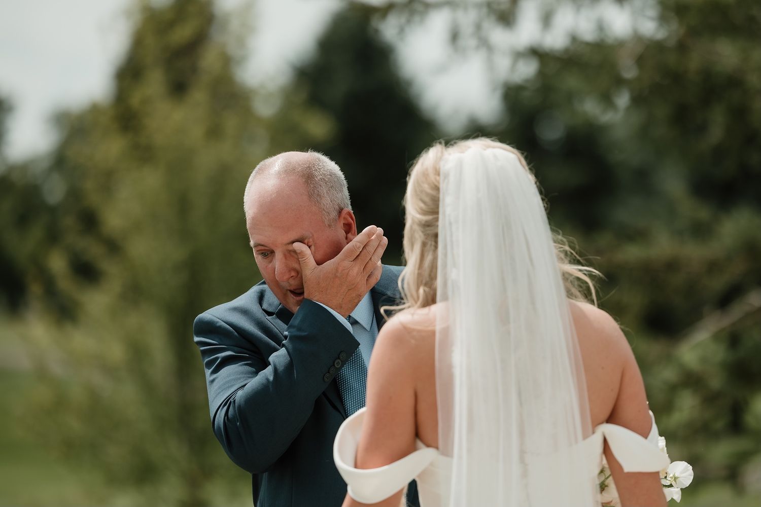 Emotional father wiping tears as he sees his daughter in her wedding dress outdoors.