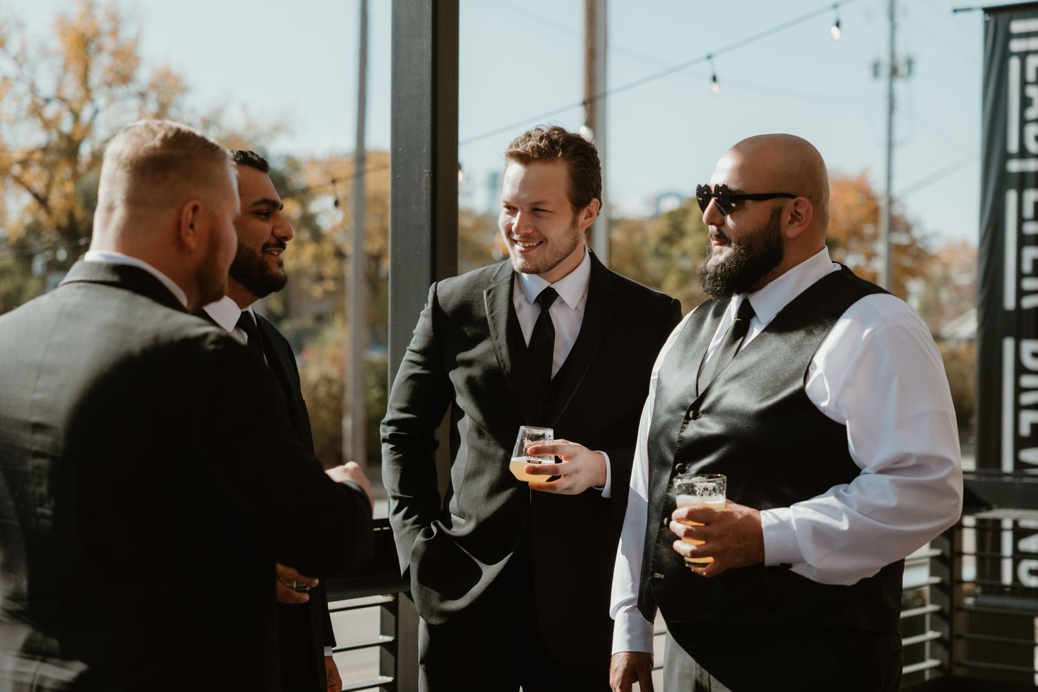 Groomsmen in formal suits and vests share drinks and conversation on a balcony during a wedding celebration.