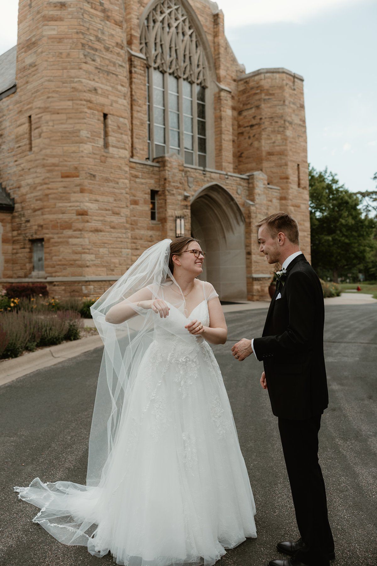 A wedding couple shares a joyful moment together outside a grand stone church with arched Gothic architecture.