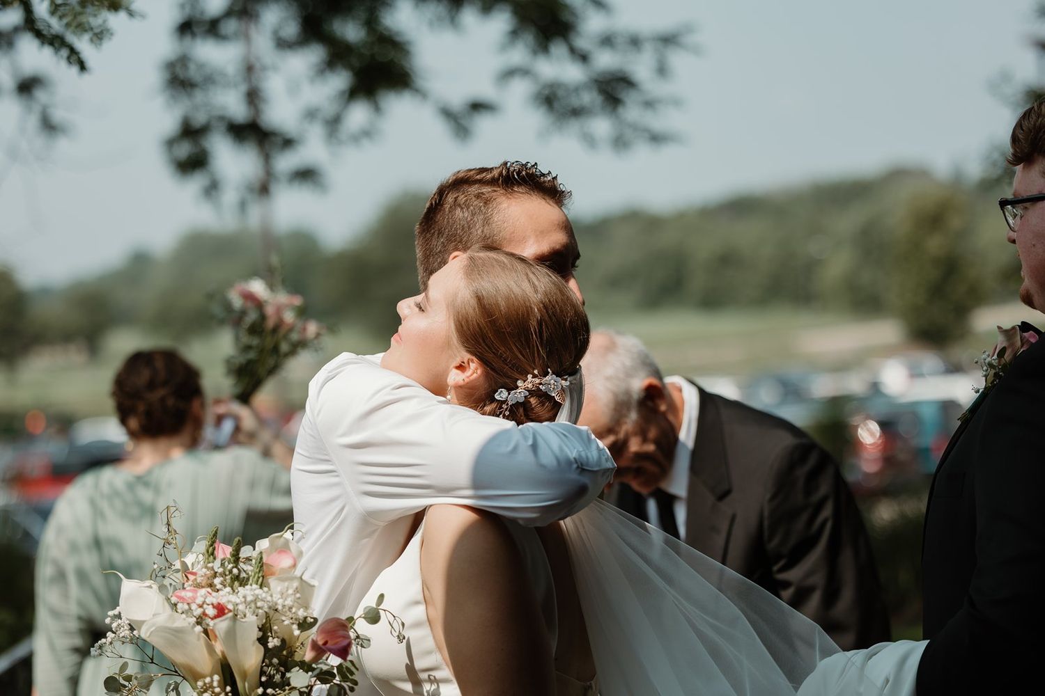 Outdoor wedding ceremony with guests seated in white chairs surrounded by natural greenery and floral arrangements.