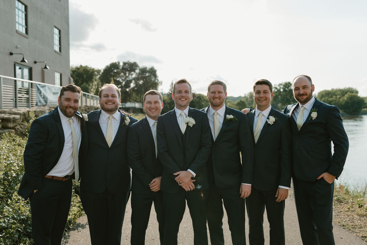 A group of groomsmen in black suits pose together outdoors for wedding photos.