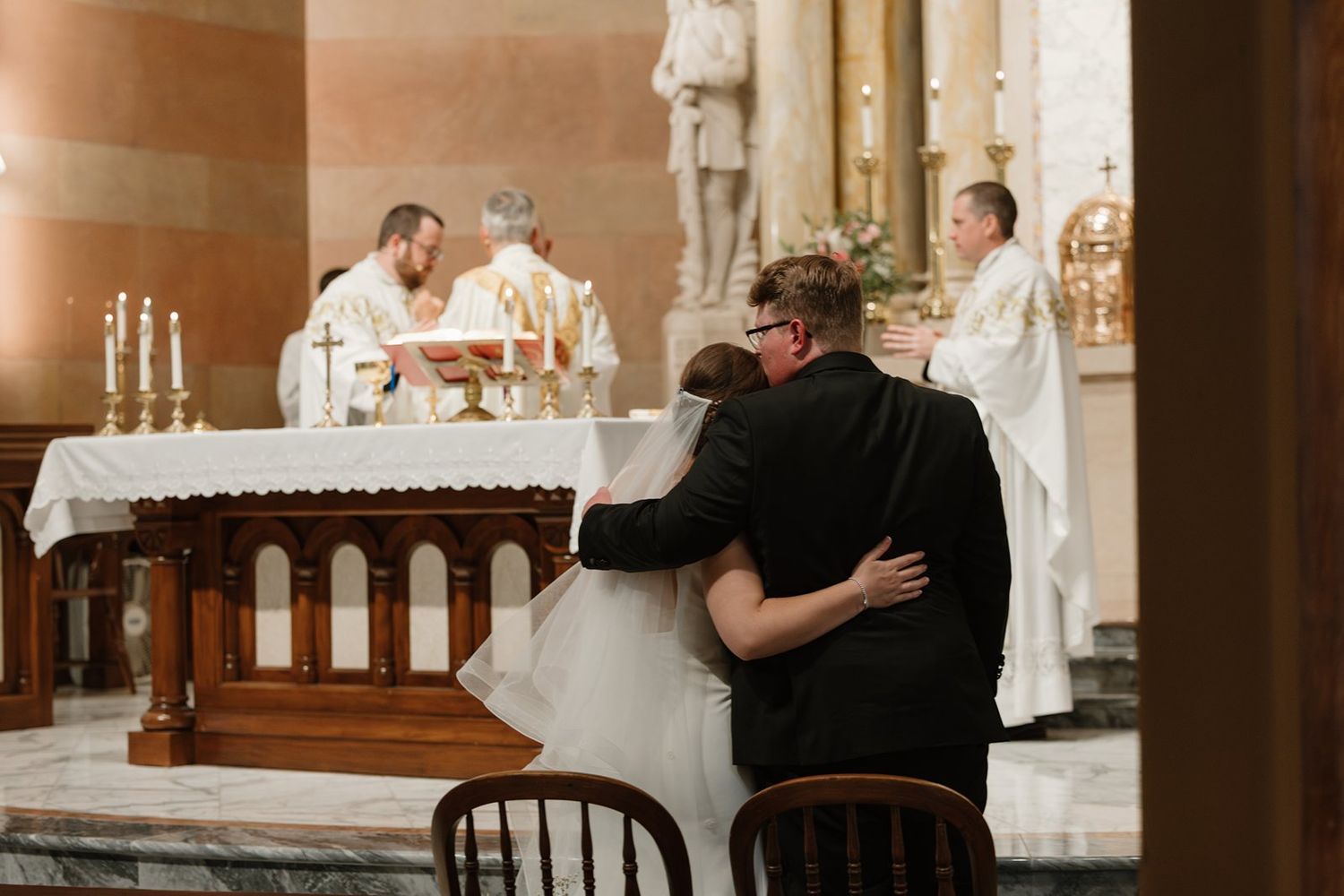 A couple embraces during their wedding ceremony inside a traditional church with white altar and wooden pews.