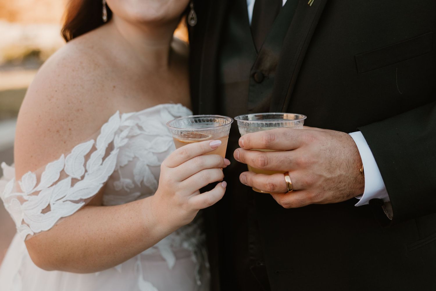 Newlyweds clink glasses together during a wedding celebration, showing detailed lace dress sleeve and formal suit.