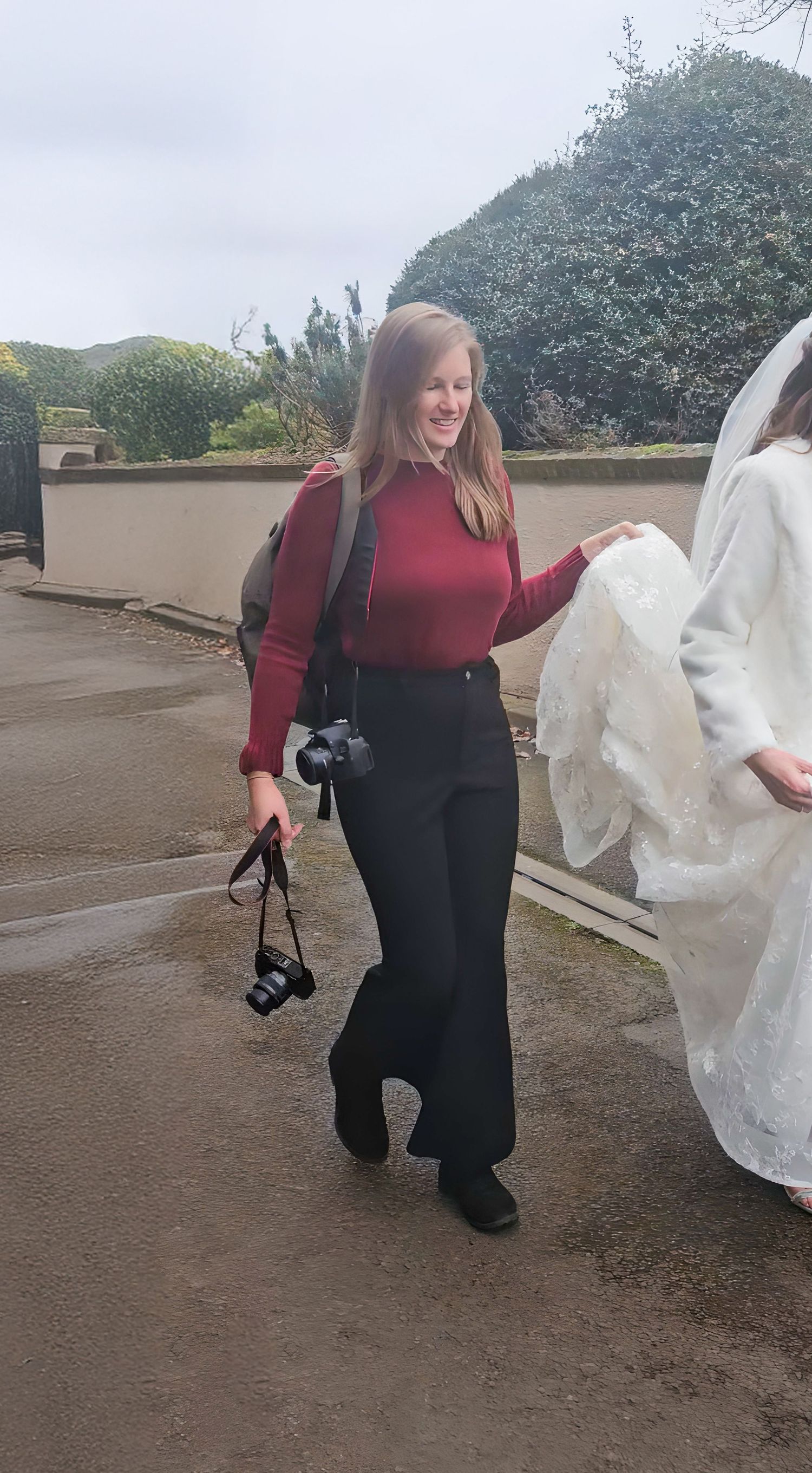 Wedding photographer in burgundy captures moments while holding camera equipment outdoors.