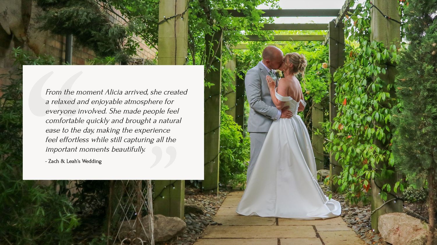 A couple in wedding attire walks through a lush garden pathway with green vines and foliage surrounding them.