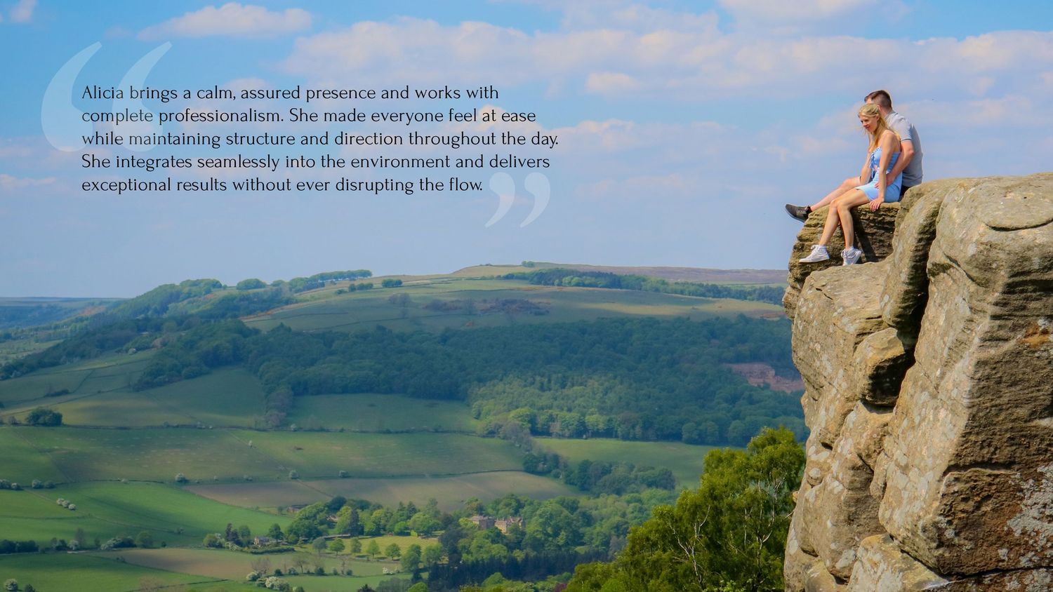 A person stands on a rocky cliff edge overlooking rolling green hills and valleys in a scenic mountain landscape.