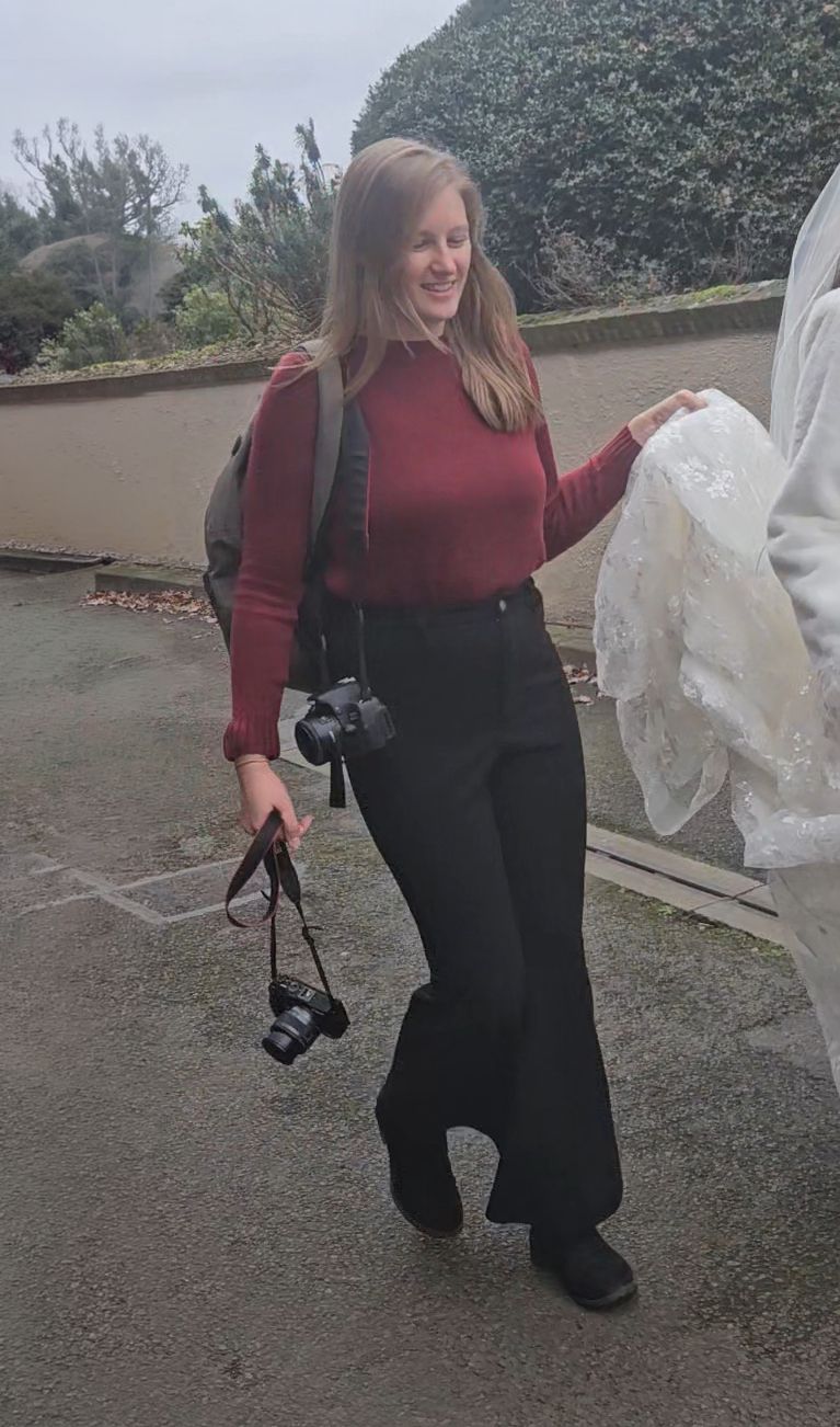 Woman in red sweater and black flare pants holding a camera outdoors, smiling while assisting at a photoshoot.