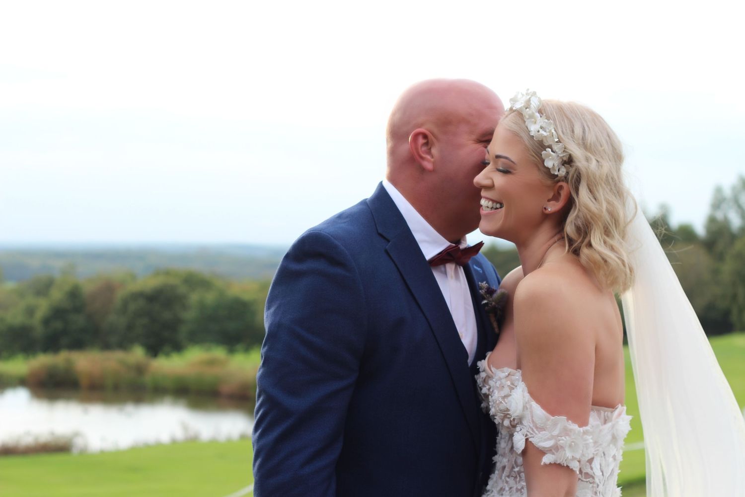 Bride and groom sharing a romantic moment outdoors, smiling together on their wedding day with a scenic lake backdrop.