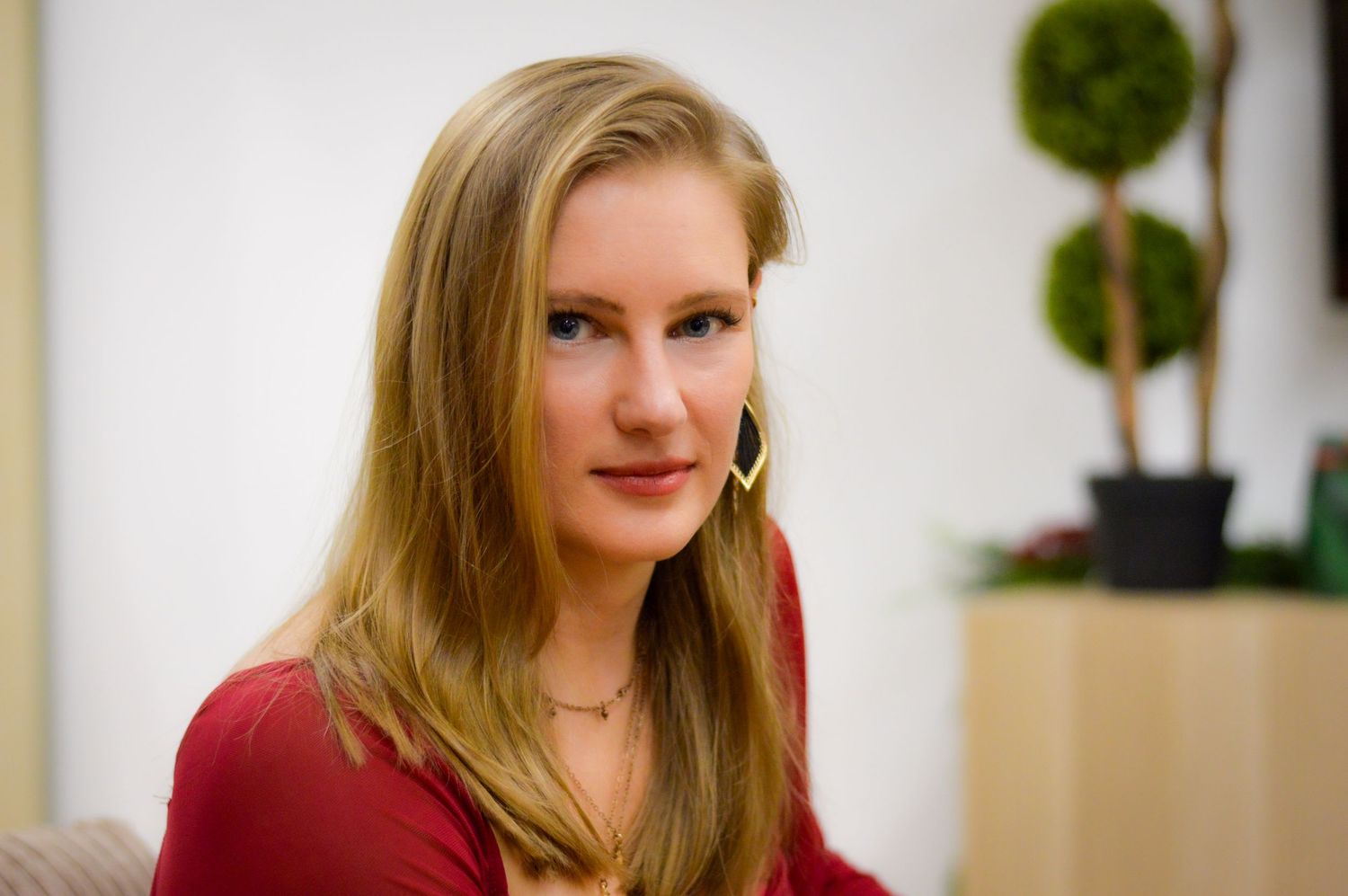 Young blonde woman wearing a red top and gold hoop earrings, posing indoors with a green topiary in background.