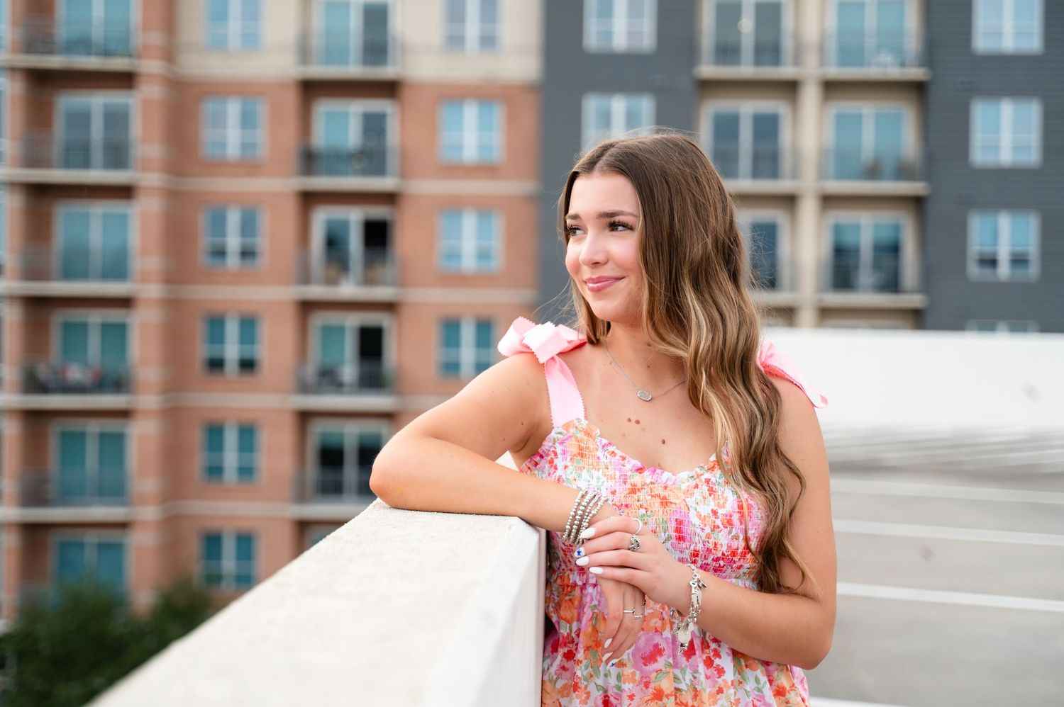 Woman in floral dress smiling while leaning on balcony with apartment building in background.