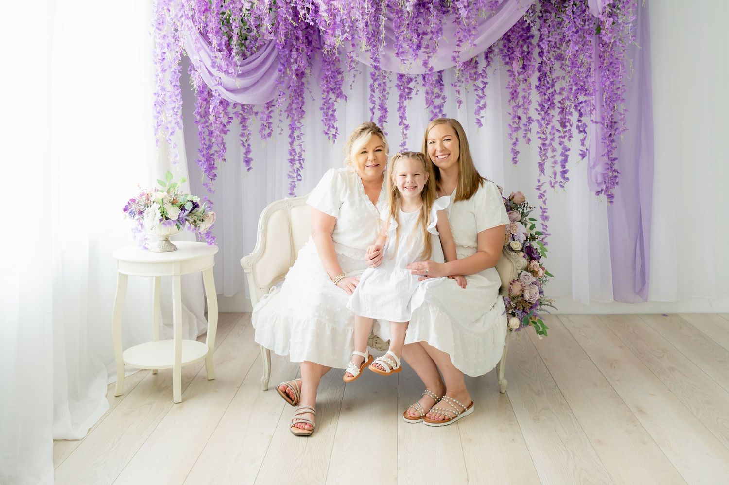 Three people in white dresses posing together against lavender floral backdrop.