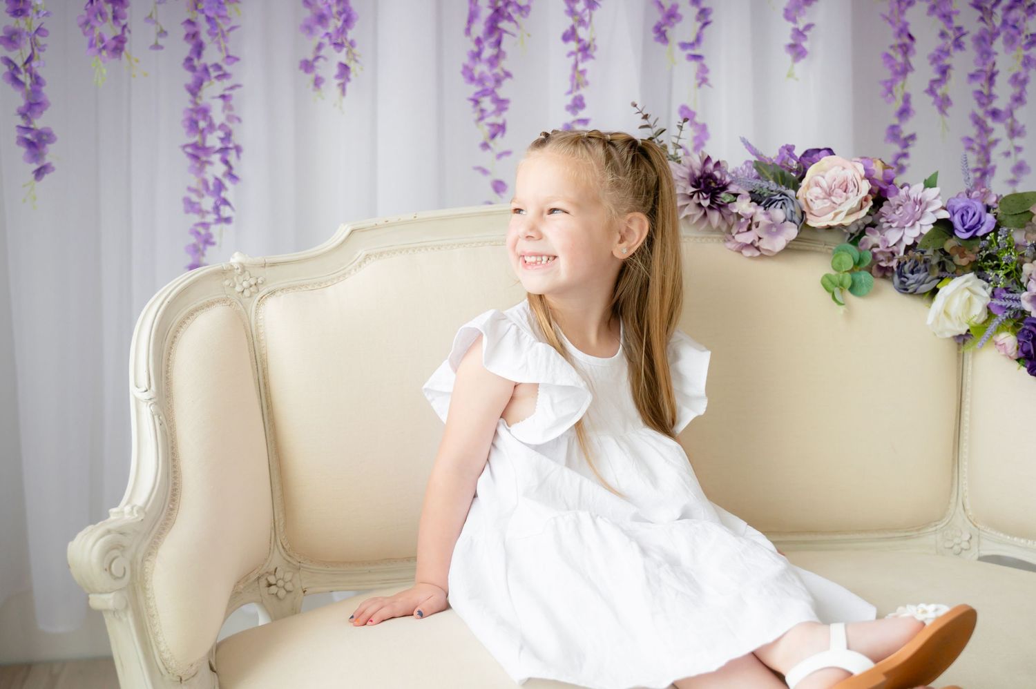 Young person in white dress sitting on vintage cream sofa with purple wisteria backdrop.