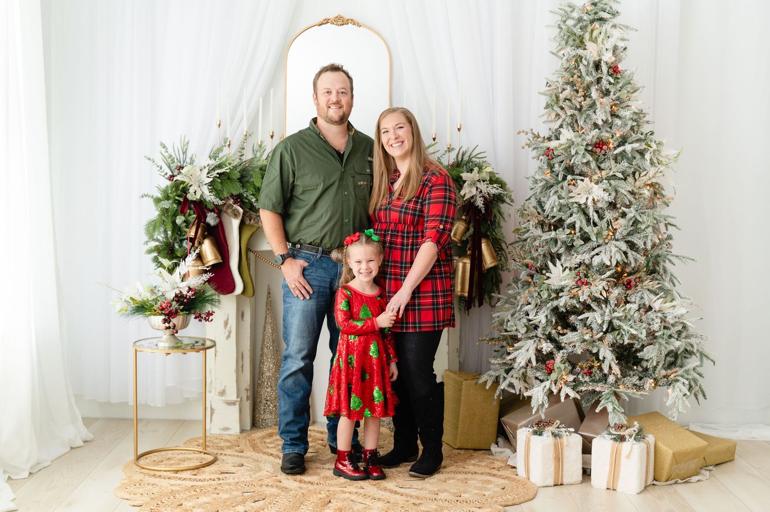 A family poses together in front of a decorated Christmas tree and white backdrop with wrapped presents and holiday decor.
