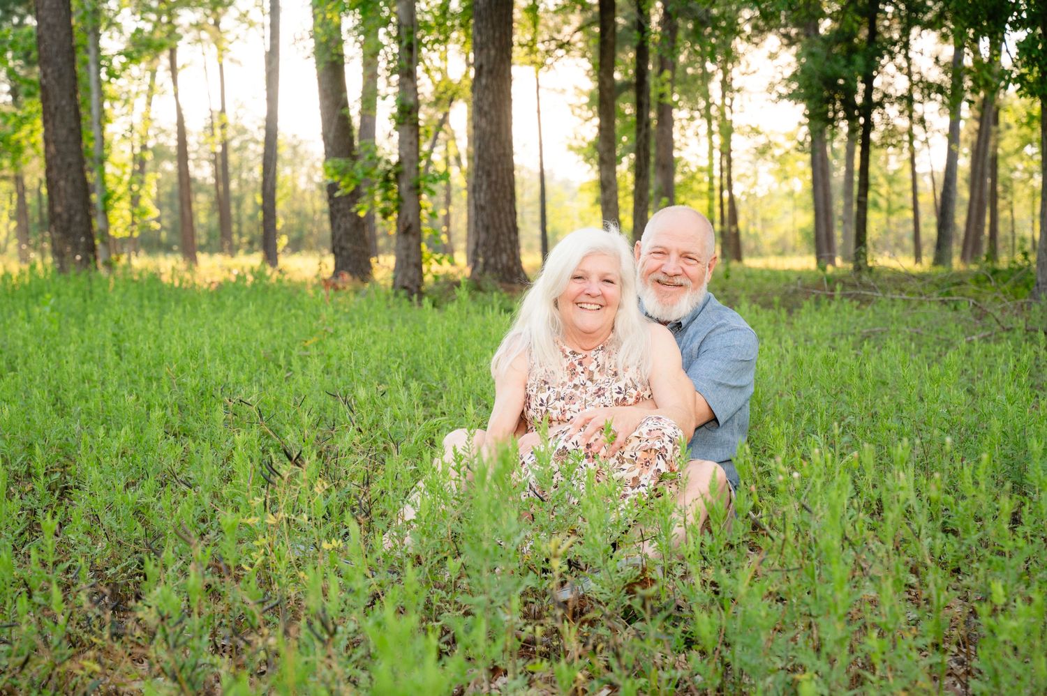 A couple sits together in a sunlit field with tall trees and wildflowers around them.